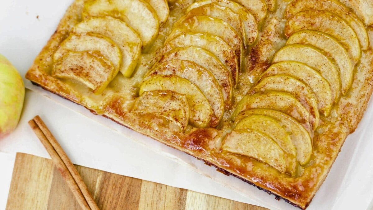 Rectangular apple tart with golden apple slices, a flaky crust, and a light glaze. Two cinnamon sticks lie nearby on a wooden surface.