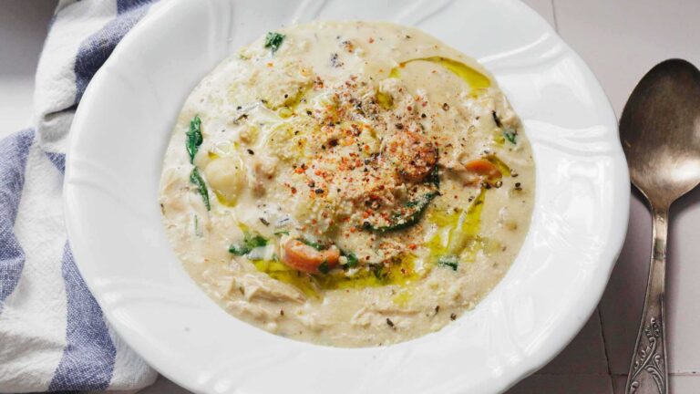 A white bowl of creamy soup with visible herbs, chunks of vegetables, and a drizzle of oil, next to a spoon and striped cloth napkin.