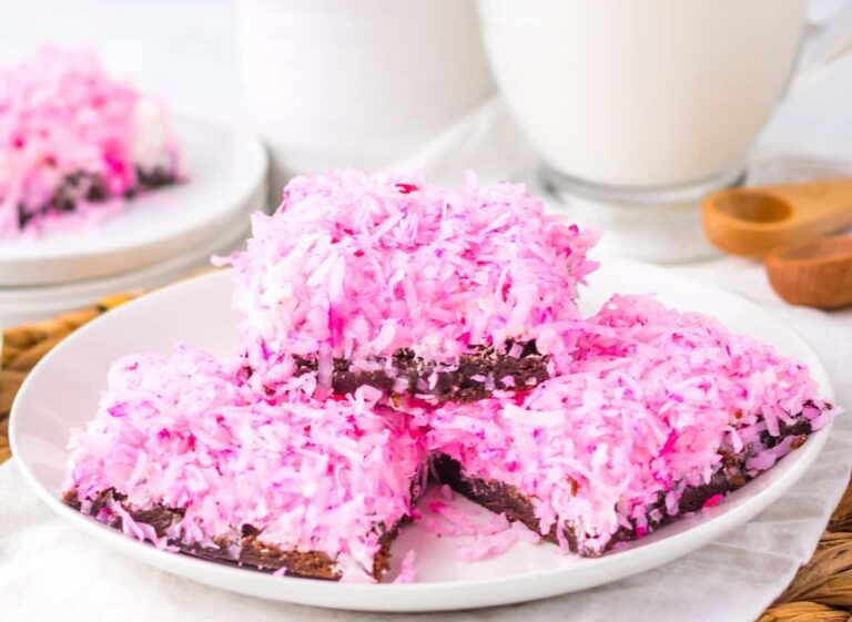 Three dessert bars topped with pink shredded coconut are stacked on a white plate, with a glass of milk and additional bars visible in the background.