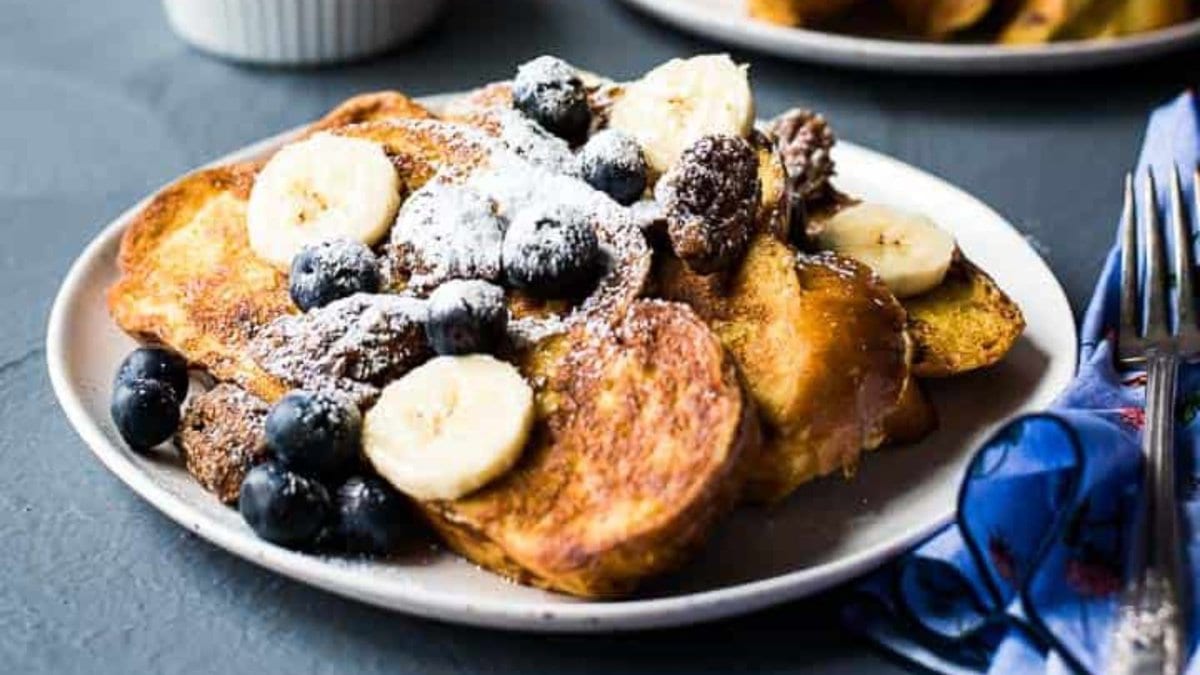 A serving of vanilla french toast next to a serving plate and bowls of candied pecans, blueberries, powdered sugar, and a banana.