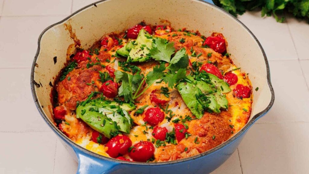 A baked casserole in a blue pot, topped with tomatoes, sliced avocado, and fresh cilantro on a white tiled surface.
