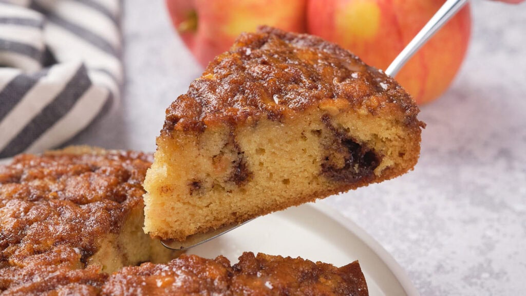 A close-up of a slice of apple cinnamon cake being lifted from the whole cake, with apples in the background.