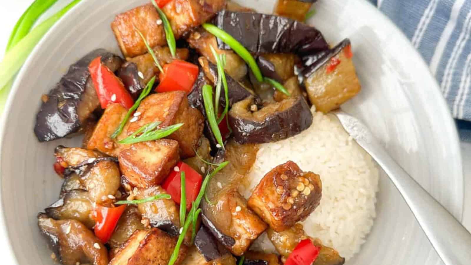 A bowl of rice topped with stir-fried eggplant, tofu, red bell peppers, and green onions. A fork is placed on the side.