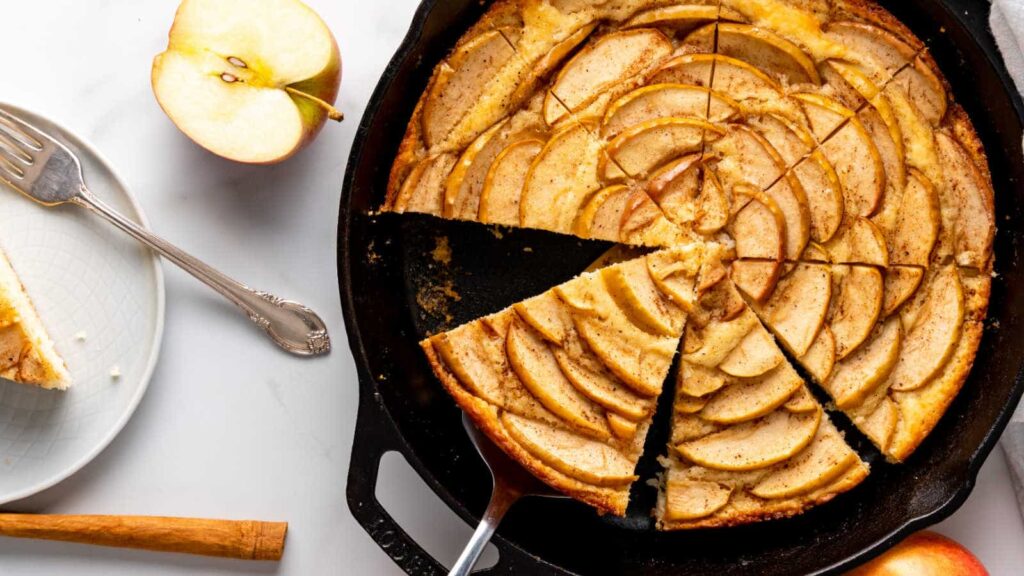 A sliced apple tart in a cast iron skillet, with a serving knife, a fork, and a plated slice next to a halved apple on a white surface.