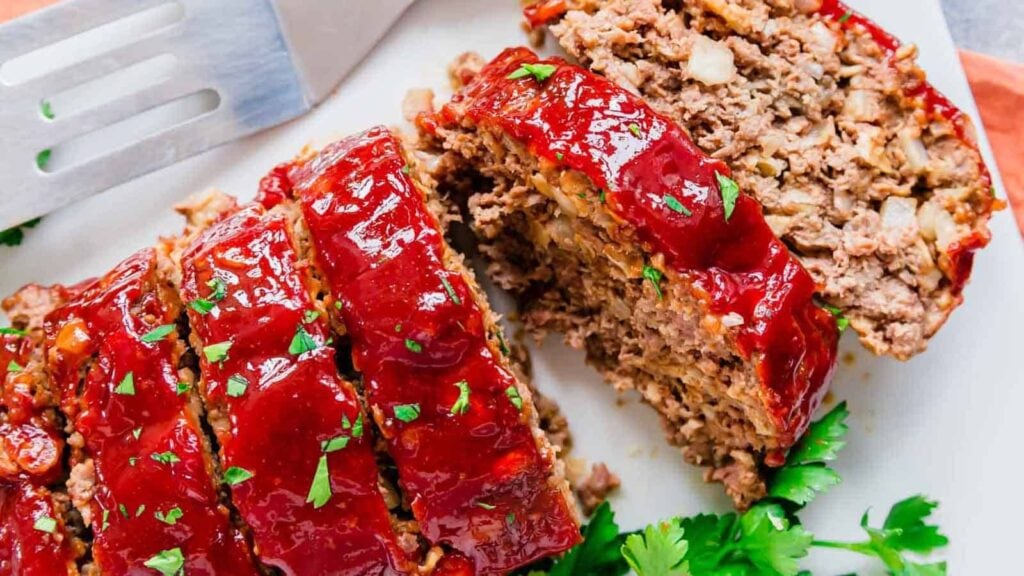 Sliced meatloaf topped with a shiny ketchup glaze, garnished with chopped herbs, served on a white platter with a spatula nearby.