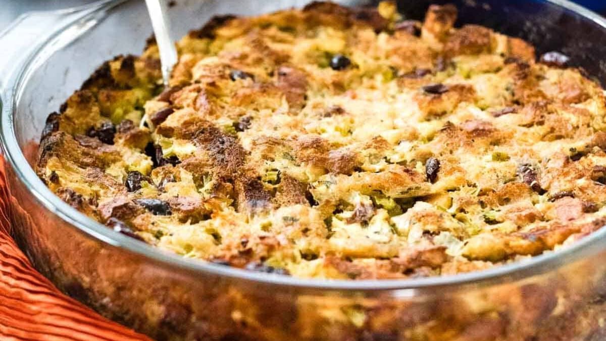 A glass baking dish filled with a baked stuffing sits on a wooden cutting board. A serving spoon is in the stuffing, and a red cloth is draped to the left, perfect for cozy fall recipes.