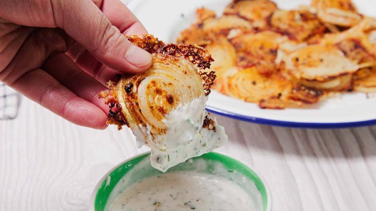 A hand dips a crispy onion slice into a bowl of creamy white dip, with more onions on a plate in the background.