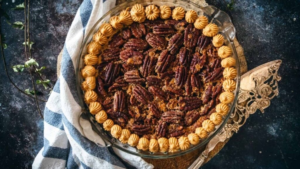 A pecan pie in a glass dish is topped with whole pecans and decorated with piped whipped cream around the edge, placed on a striped cloth.