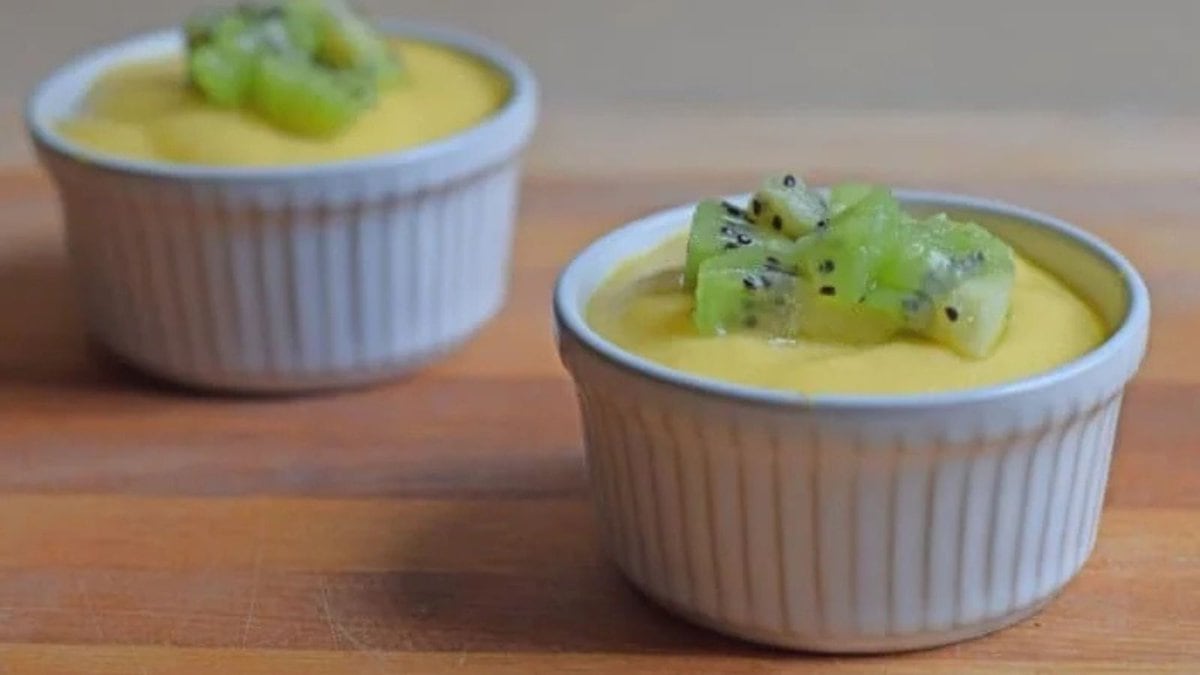 An overhead shot shows two ramekins with mango custard topped with chopped kiwi on a wooden cutting board.