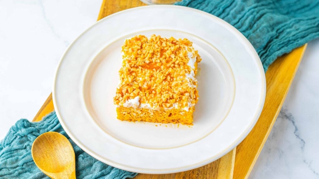A square piece of cake with white frosting and chopped nuts on top, served on a white plate with a wooden spoon beside it.