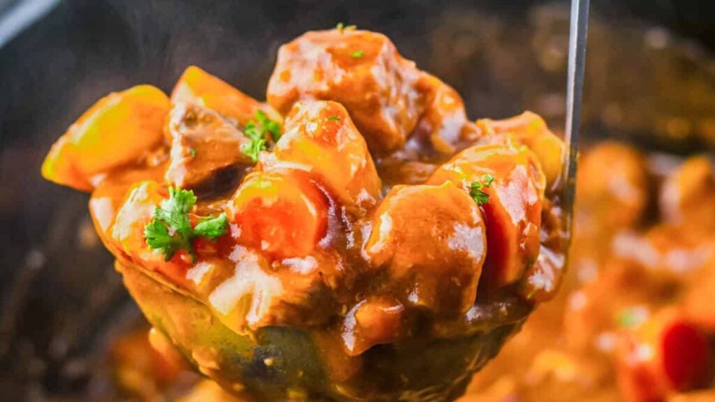 A ladle filled with thick beef stew containing chunks of meat, carrots, potatoes, and garnished with parsley.