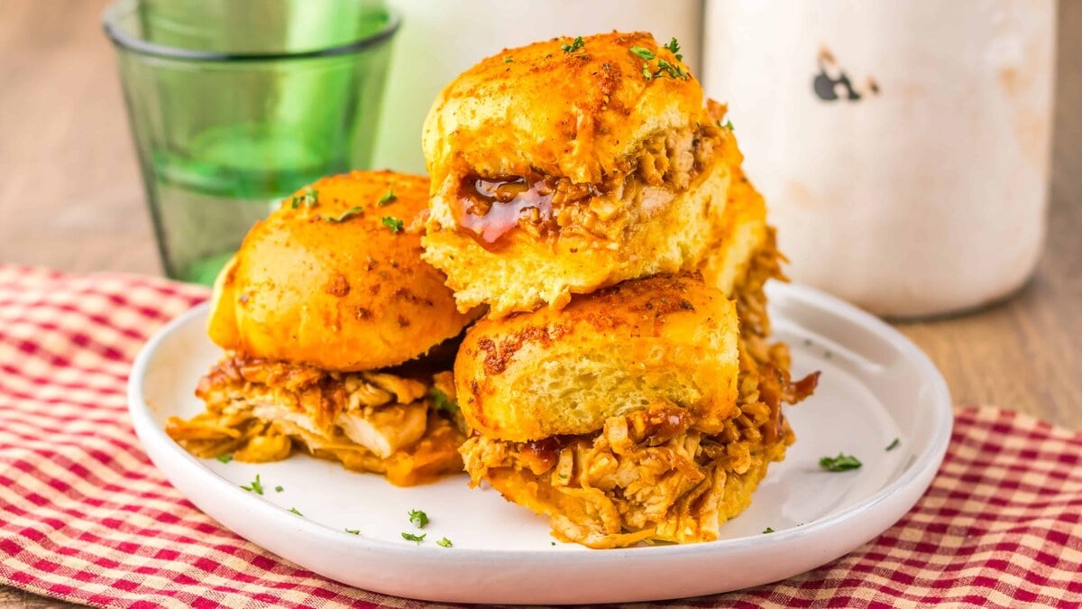 Three barbecue chicken sliders on a white plate, topped with herbs, set on a red checkered cloth with a green glass and white container in the background.