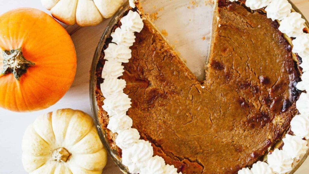 A pumpkin pie with a missing slice sits surrounded by three small pumpkins, topped with whipped cream along the edge.