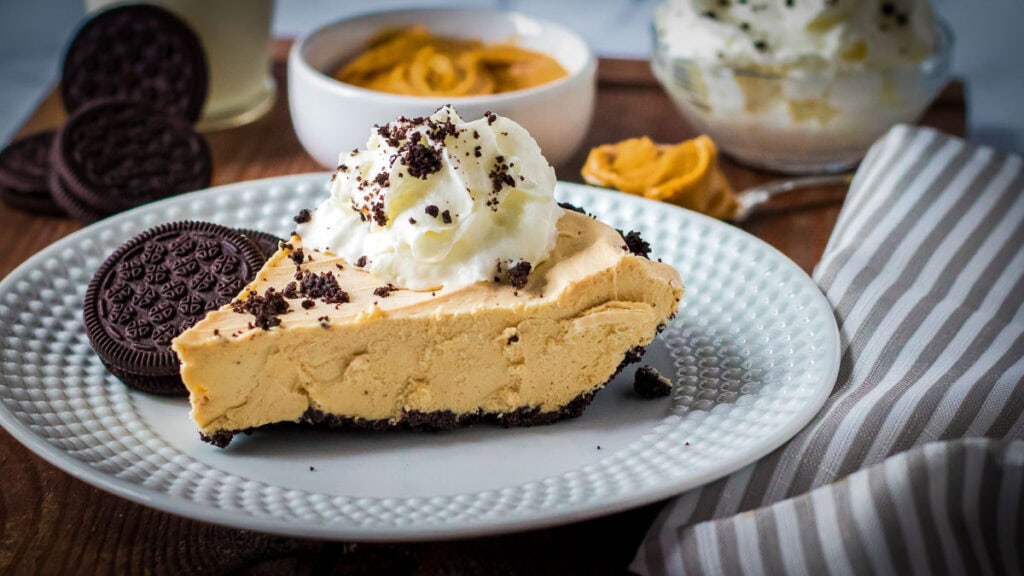 A slice of peanut butter pie with a cookie crust, topped with whipped cream and cookie crumbs, served with chocolate sandwich cookies on a white plate.