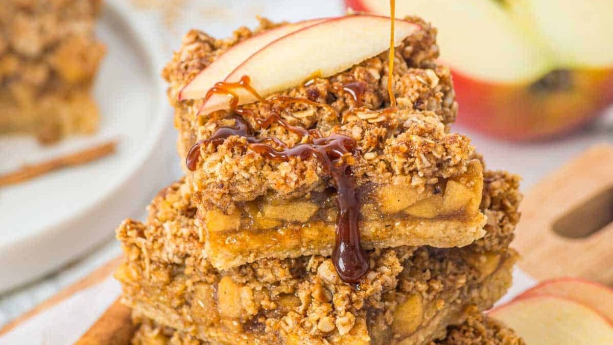 Three stacked apple crumble bars topped with a fresh apple slice, drizzled with caramel sauce.