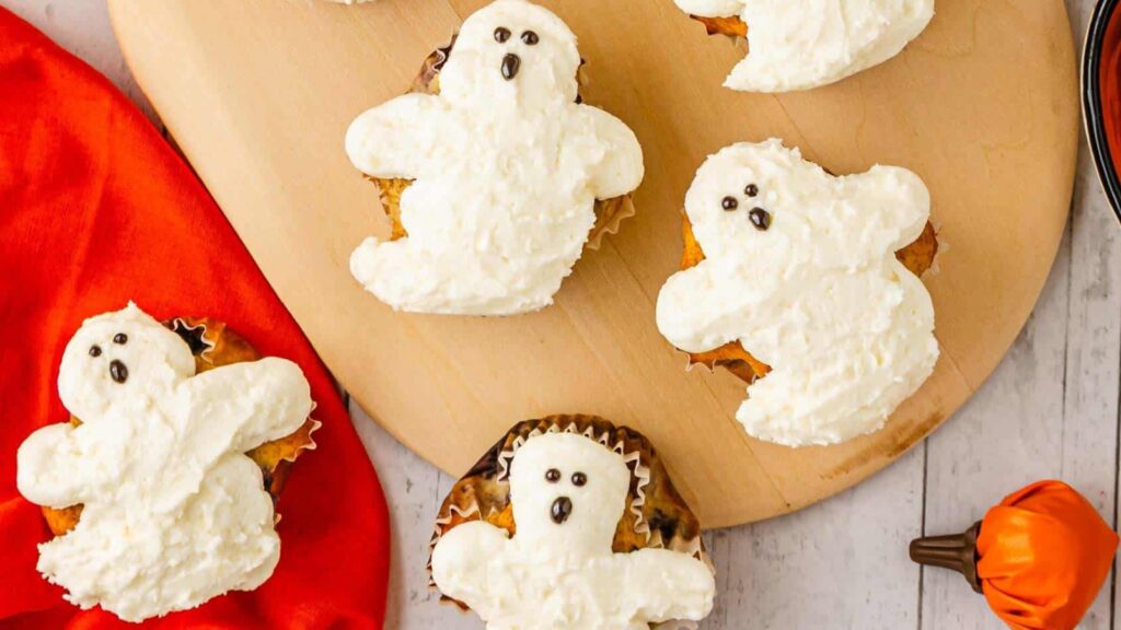 Cupcakes decorated with white frosting shaped like ghosts, each with small chocolate eyes and mouth, arranged on a wooden board.