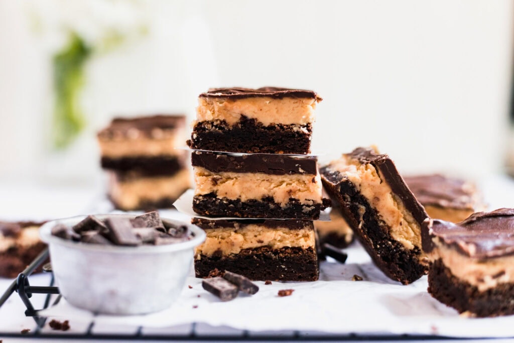 Three chocolate and peanut butter layered dessert bars are stacked on a cooling rack, with more bars and a small metal cup filled with chocolate chunks nearby.