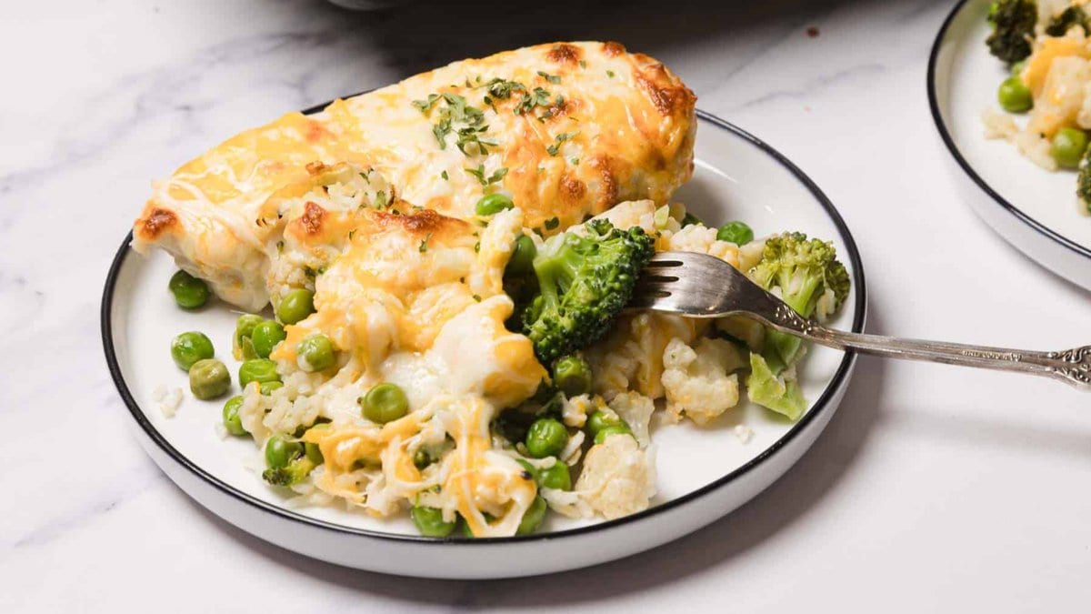 A plate with baked chicken breast topped with melted cheese, served with peas, broccoli, cauliflower, and rice. A fork rests on the plate.