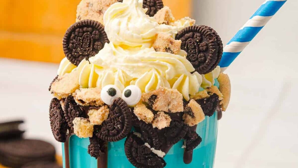A blue cup filled with a milkshake, topped with whipped cream, mini chocolate sandwich cookies, cookie pieces, candy eyes, and a blue striped straw.