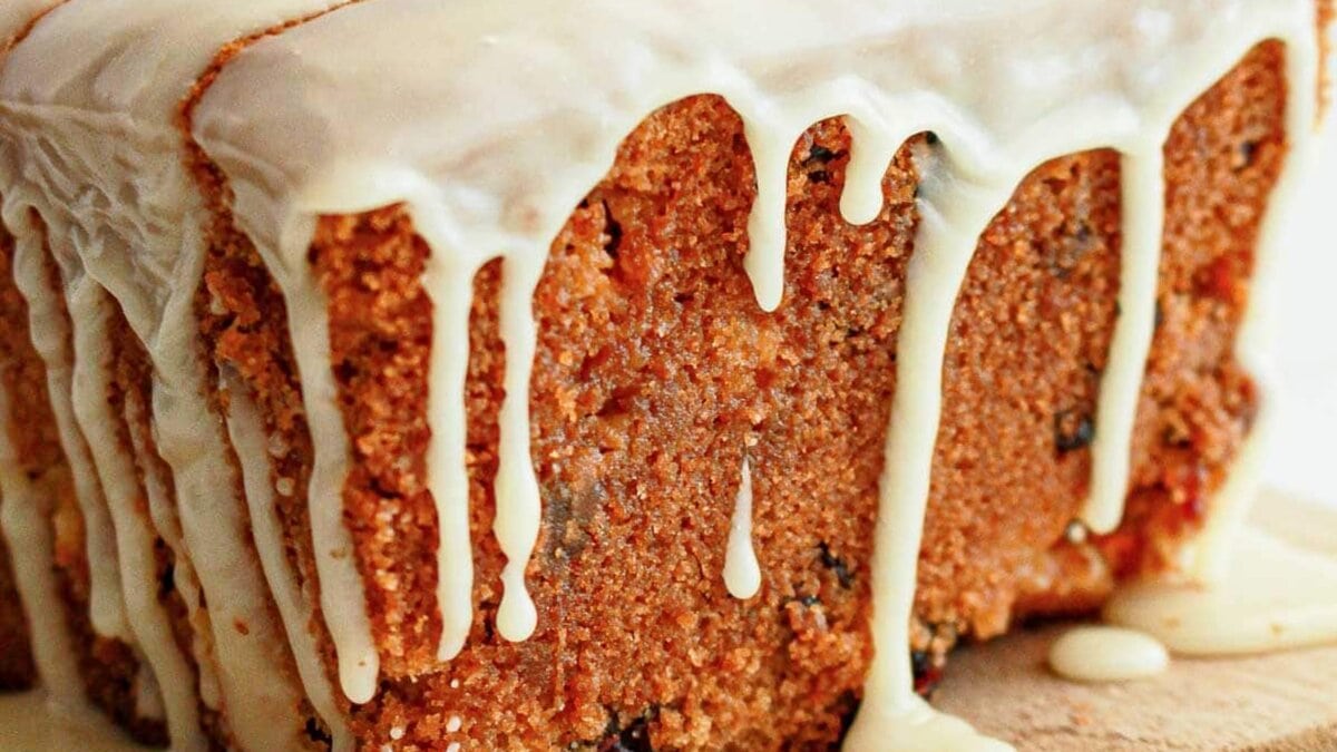 A loaf cake with white icing drips on a wooden board, next to two forks and some dried fruit pieces.