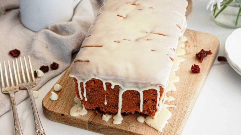 A loaf cake with white icing drips sits on a wooden board, surrounded by forks and small dried fruit pieces on a white table.
