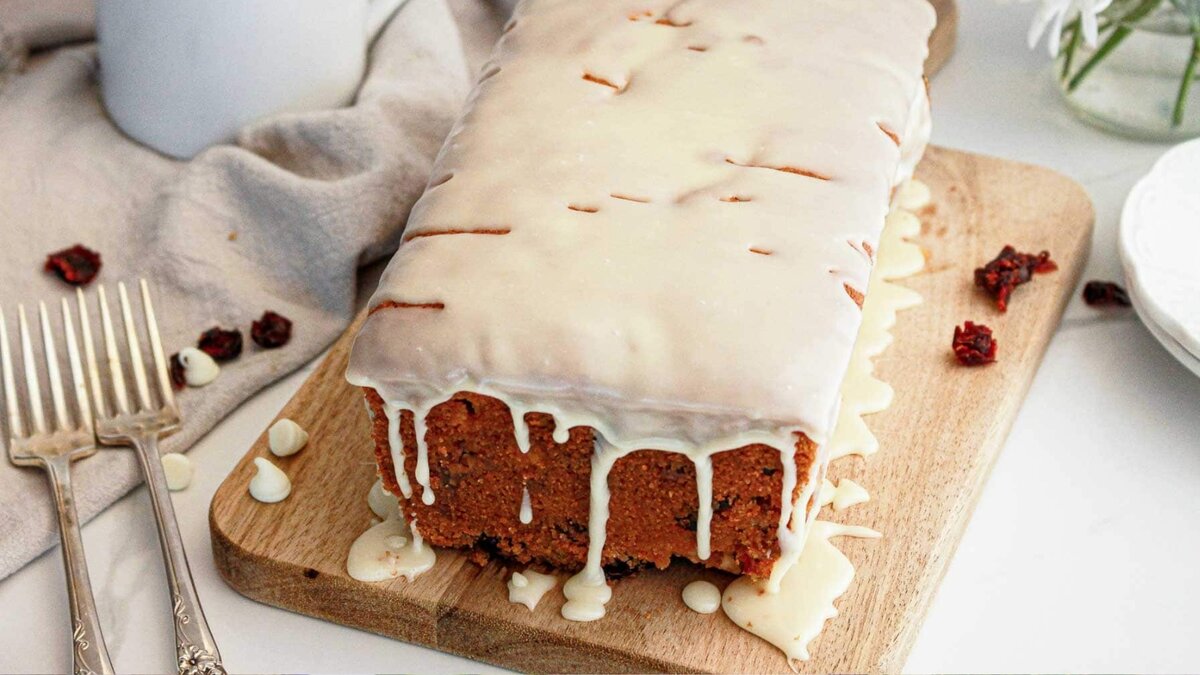 A loaf cake with white icing drips on a wooden board, next to two forks and some dried fruit pieces.