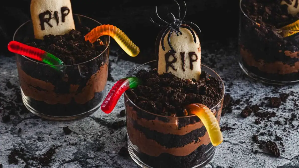 Three clear glasses filled with layers of brown mousse, topped with crumbled cookies, and decorated with gummy worms and cookie tombstones labeled 