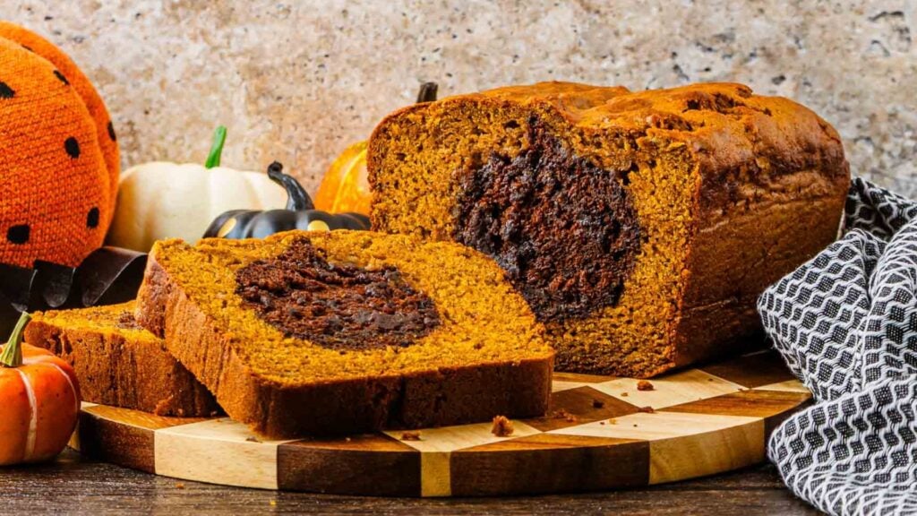 A loaf of pumpkin bread with a chocolate swirl center sits on a wooden board; two slices are cut, surrounded by small decorative pumpkins.