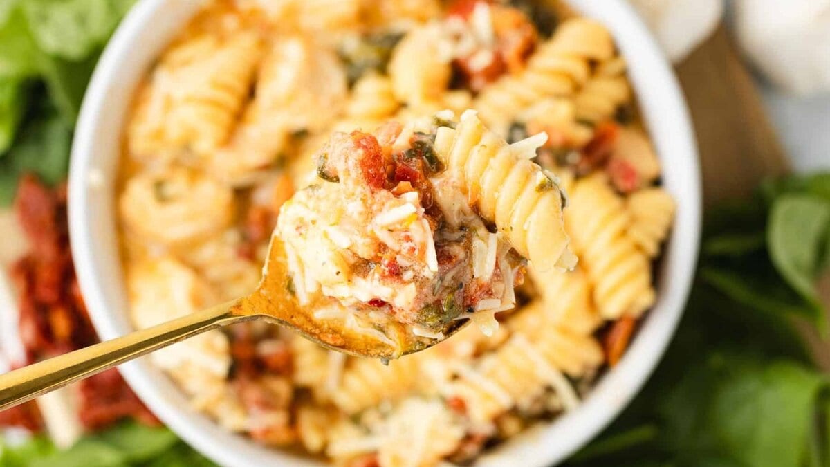 A close-up of a fork lifting a bite of rotini pasta with tomato, cheese, and herbs from a white bowl of pasta.