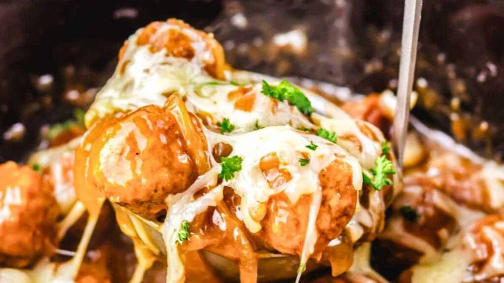 A close-up of meatballs topped with melted cheese and chopped parsley, covered in gravy, being served with a ladle.