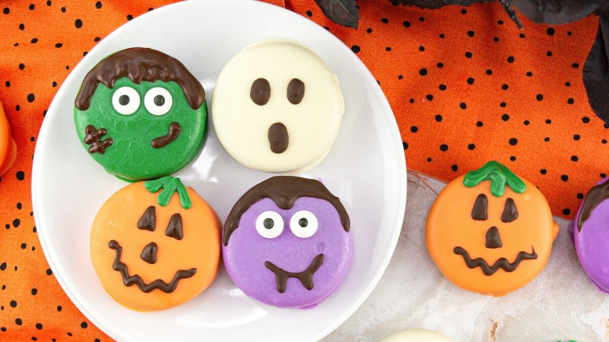 A plate of decorated Halloween Oreo cookies sits on a polka dot orange cloth. The cookies are decorated like jack-o'-lanterns, a ghost, Frankenstein and vampire, each covered in colorful icing.