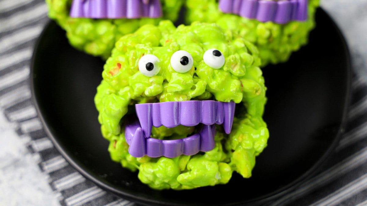 A close-up of halloween green popcorn treats with monster faces with candy eyes and purple teeth, placed on a black plate. The background shows a striped cloth.