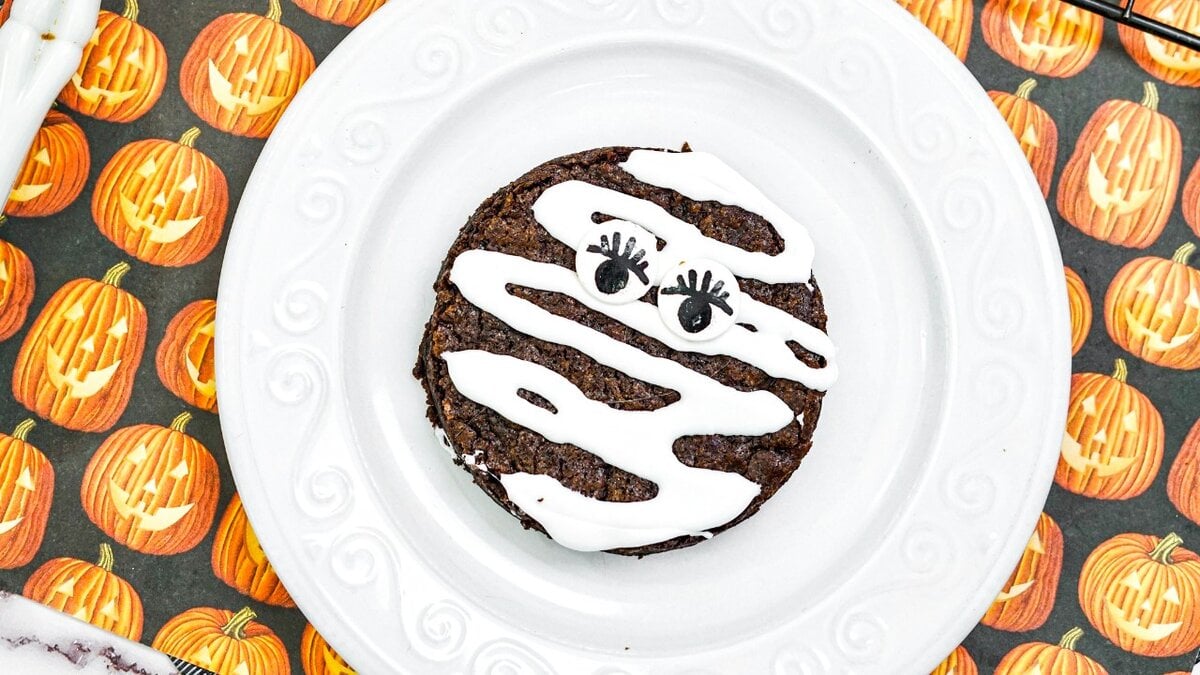 A chocolate cookie with white icing designed to resemble a mummy, featuring two candy eyes on top. The cookie is placed on a white plate over a pumpkin-themed tablecloth.