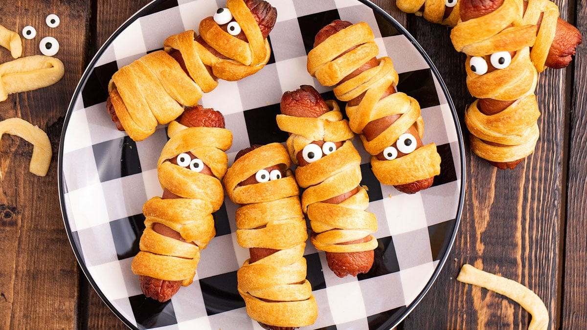 Mummy Halloween hot dogs wrapped in crescent dough with mustard eyes on a parchment-lined baking sheet.