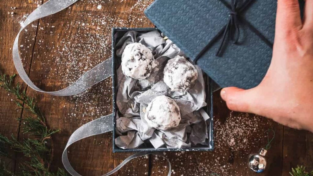 A hand lifts the lid of a textured blue gift box containing three powdered sugar-covered cookies on crumpled paper, set on a wooden table with ribbon and scattered sugar.