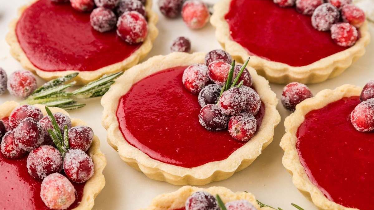 Mini cranberry tarts with smooth red filling, topped with sugared cranberries and rosemary sprigs, arranged on a light surface.