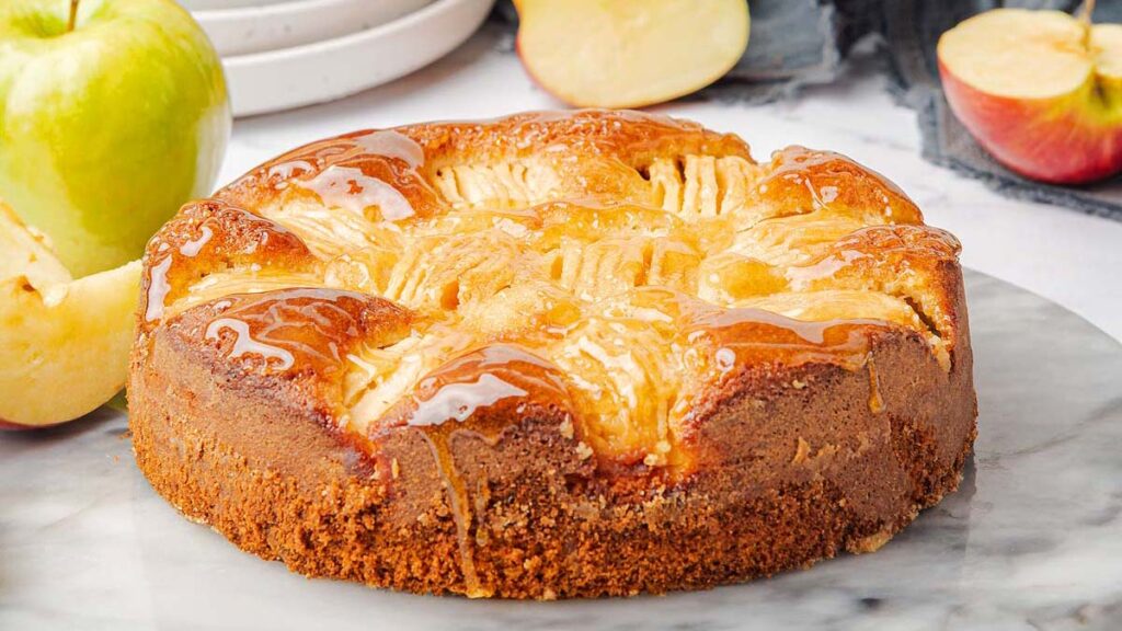 A round apple cake with a golden-brown crust and glazed top sits on a marble surface, with fresh apple slices in the background.