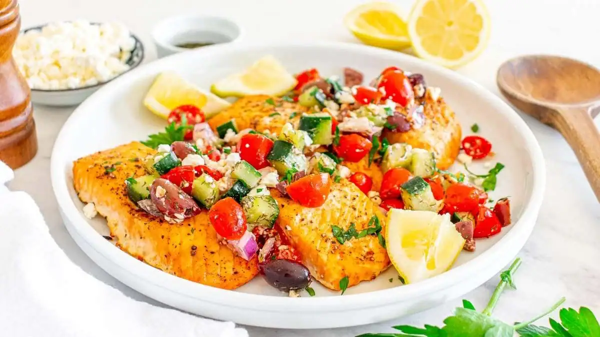 Grilled salmon fillets topped with a Mediterranean salad of cherry tomatoes, cucumber, olives, red onion, and feta, garnished with lemon wedges on a white plate.