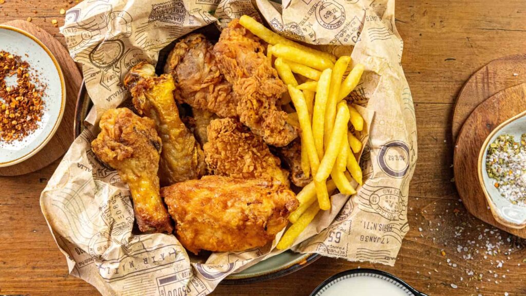 A basket lined with printed paper holding fried chicken pieces and French fries, placed on a wooden table with seasoning plates nearby.