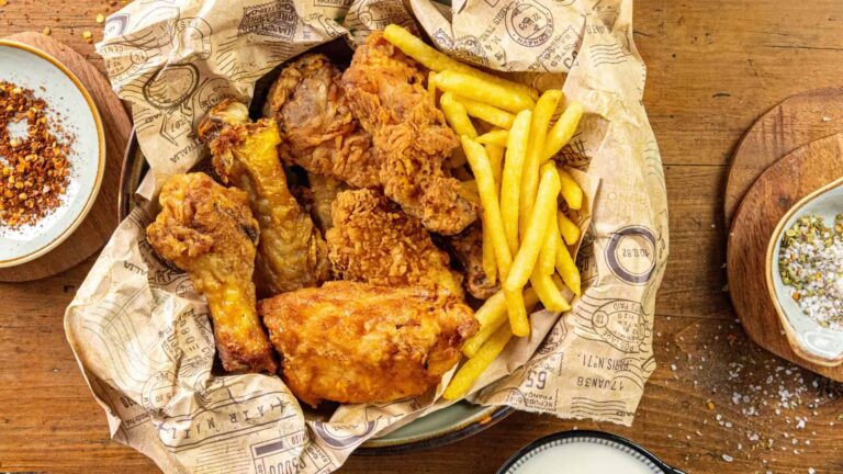 A basket lined with printed paper holding fried chicken pieces and French fries, placed on a wooden table with seasoning plates nearby.