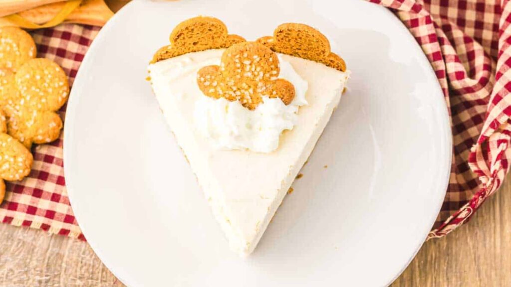 A slice of cheesecake topped with whipped cream and two gingerbread cookies sits on a white plate beside a red checkered napkin.