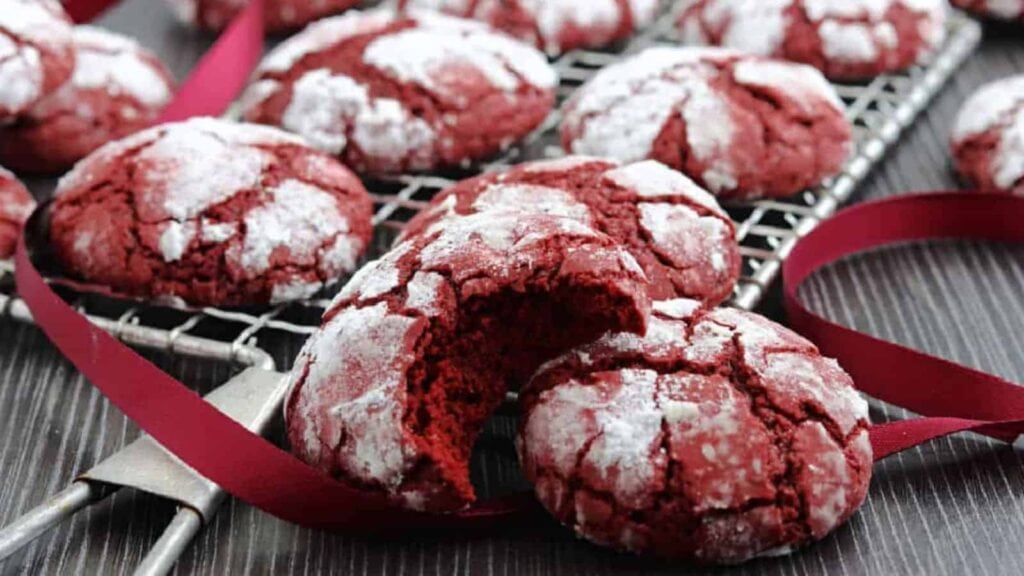 Red velvet crinkle cookies covered with powdered sugar are displayed on a cooling rack, with one cookie partially eaten and a red ribbon looped around the rack.
