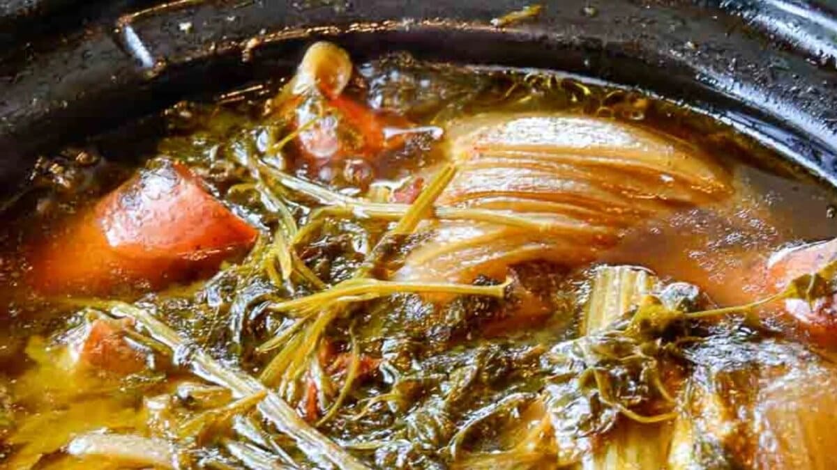 Close-up of a stew featuring leafy greens, carrots, onions, and broth in a dark pot.