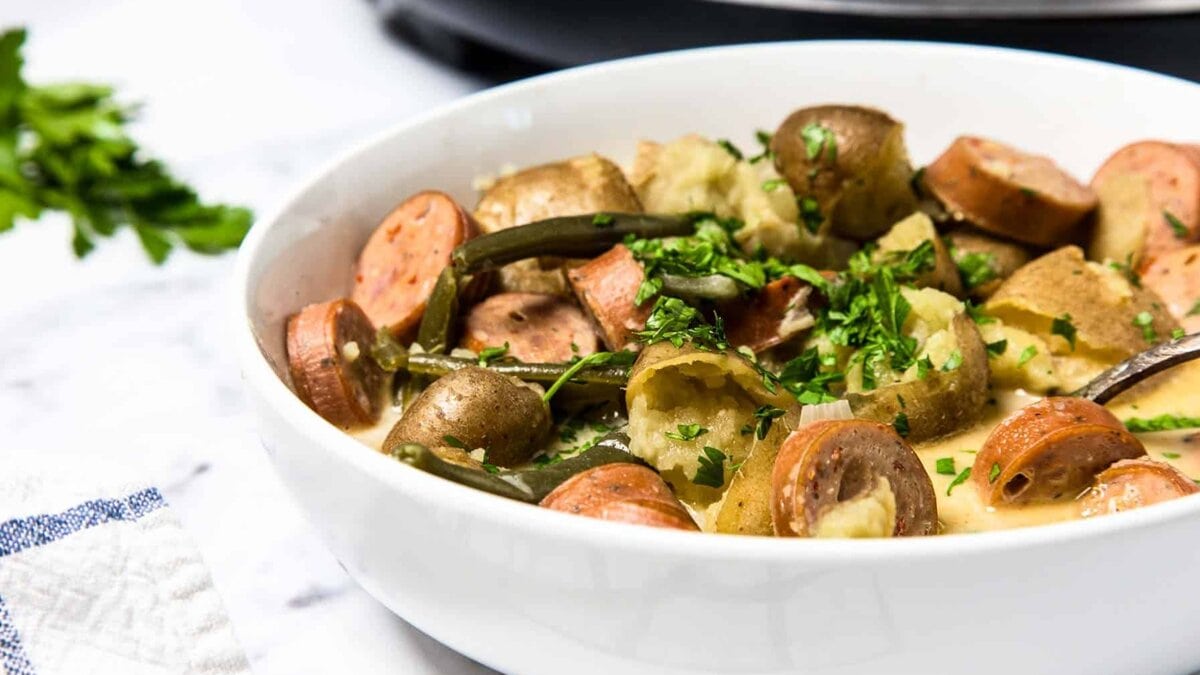 A white bowl filled with sliced sausage, potatoes, green beans, and chopped parsley on a light surface.
