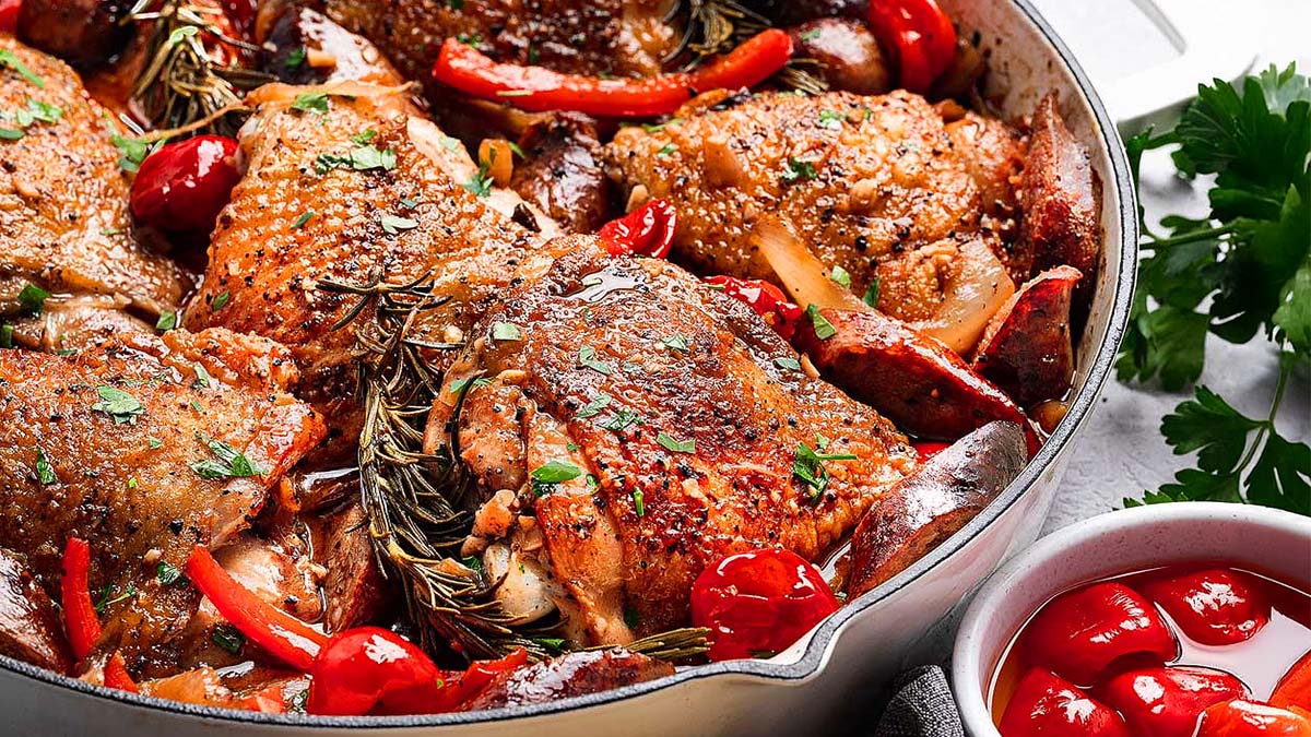 A pan of roasted chicken thighs with herbs, red bell peppers, and cherry tomatoes, garnished with chopped parsley.