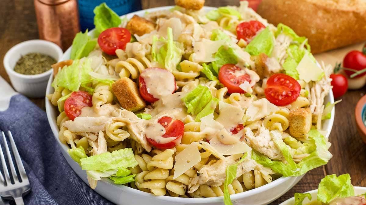 A bowl of pasta salad with rotini noodles, lettuce, cherry tomatoes, shredded chicken, croutons, shaved parmesan, and creamy dressing.