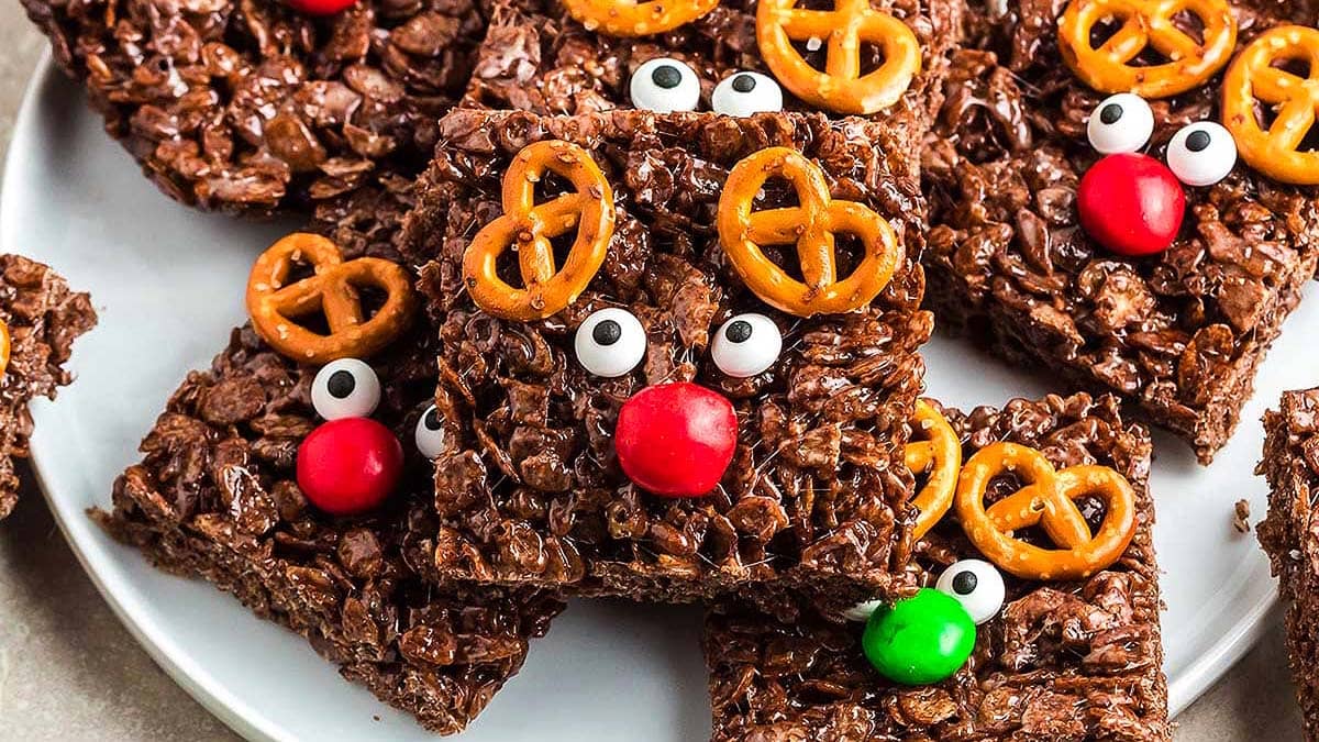 Chocolate rice krispie treats decorated with pretzel antlers, candy eyes, and red or green candy noses to resemble reindeer, arranged on a white plate.