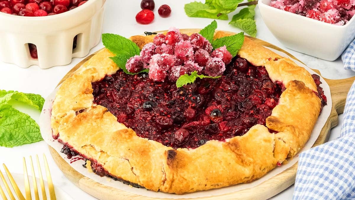 A rustic berry galette topped with sugared cranberries and mint leaves, surrounded by bowls of fresh berries and mint on a white surface.
