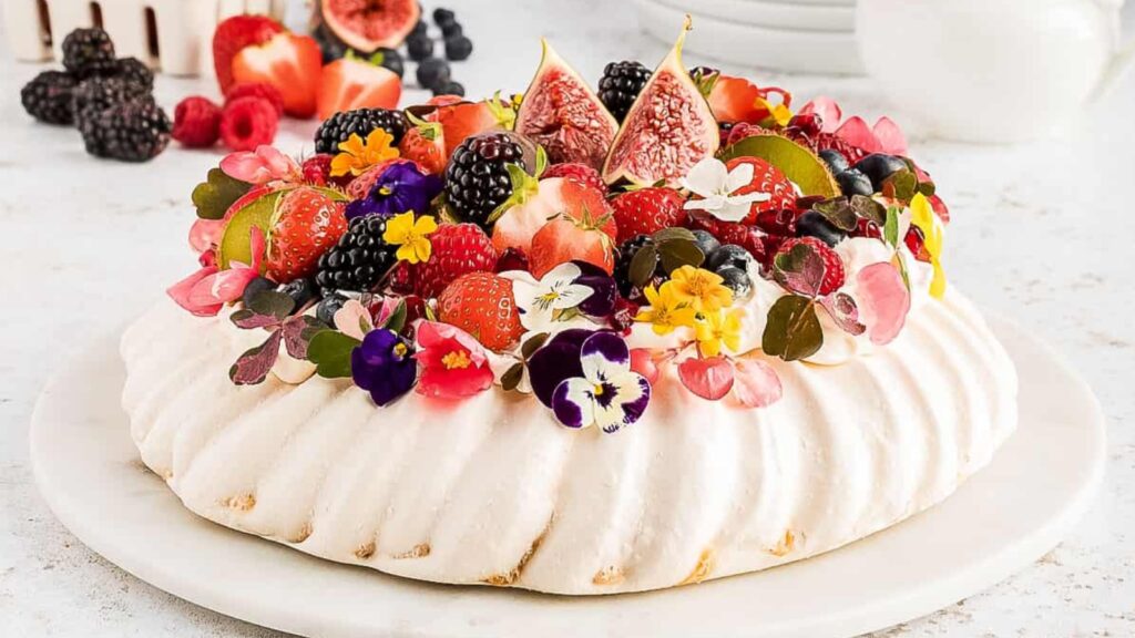 A pavlova topped with whipped cream, mixed berries, figs, and edible flowers sits on a white plate.
