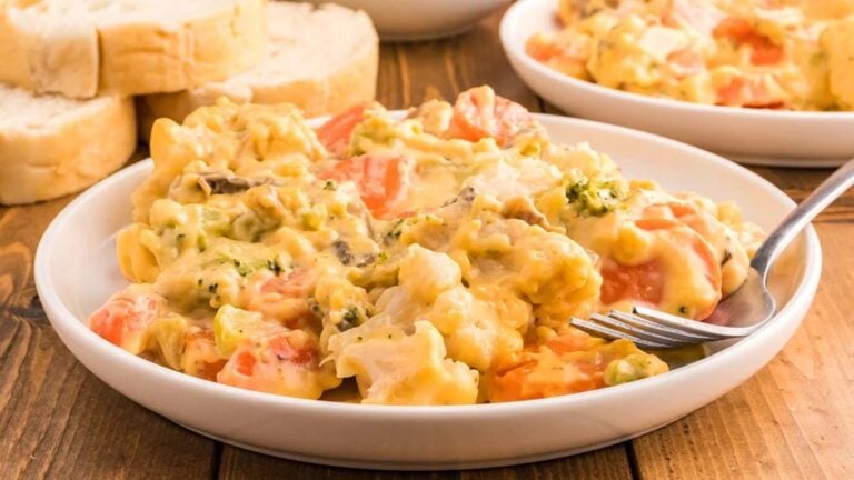 A plate of creamy vegetable casserole with visible broccoli, carrots, and cauliflower, served next to slices of bread on a wooden table.